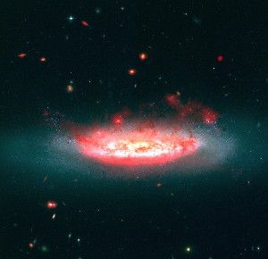 A composite image shows the galaxy NGC 4522 in the Virgo Cluster, the nearest large cluster of galaxies to our own local group of galaxies, and the “wake” of gas and dust being blown from the galaxy. The galaxy appears blue in the Hubble Space Telescope image in visible light. The superimposed red image is from Spitzer data and shows emissions from dust that traces molecular hydrogen. In the image, the galaxy is moving down and into the plane of the photo.